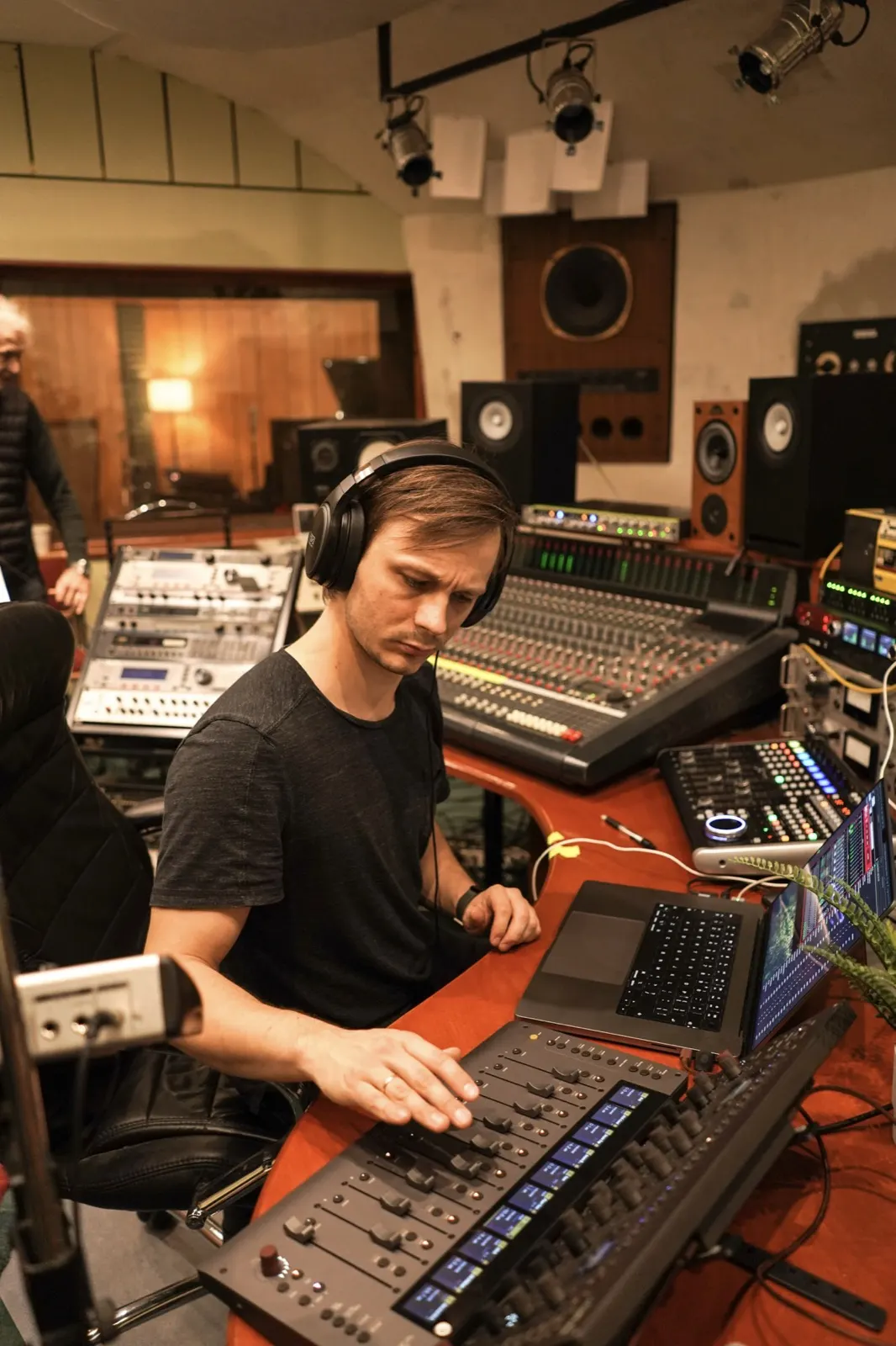 Toms at a studio desk with headphones on, riding faders on a control surface